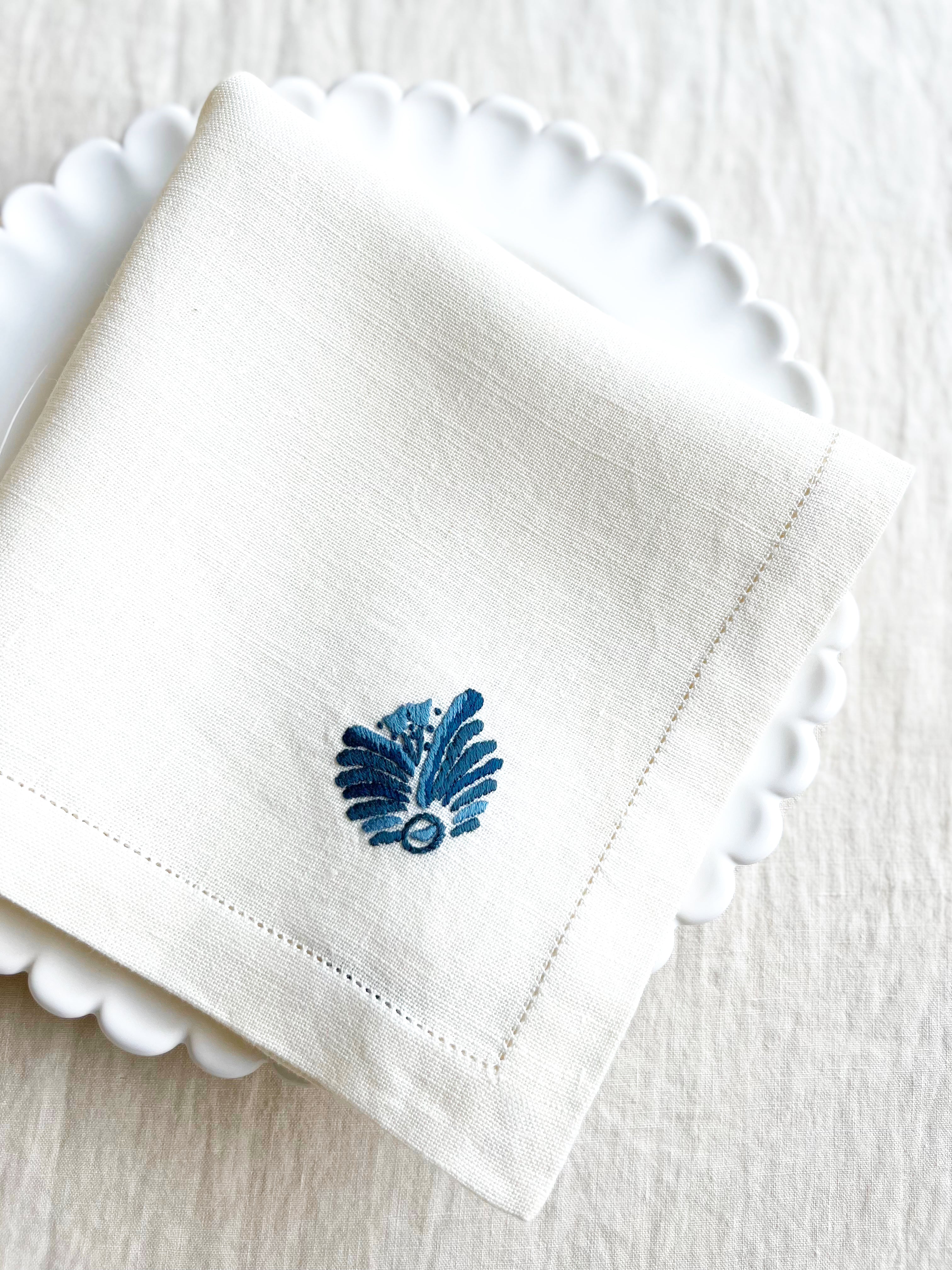 white hand embroidered linen napkins with blue palm frond in corner 16 inches square folded on plate