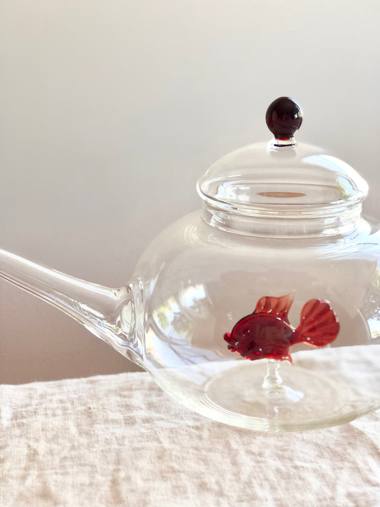 clear hand blown glass teapot with red glass fish in bottom on white table